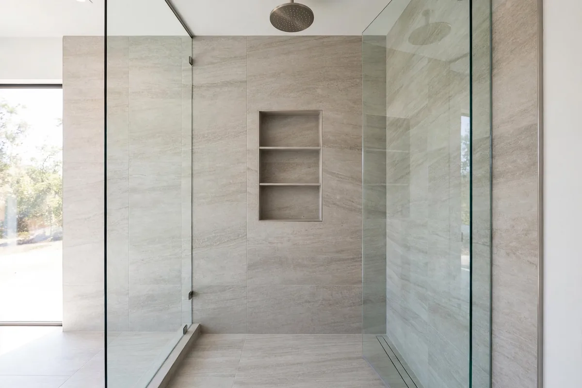 Curbless walk-in shower with frameless glass panels, rainfall showerhead and recessed tiled niche in large-format stone tile